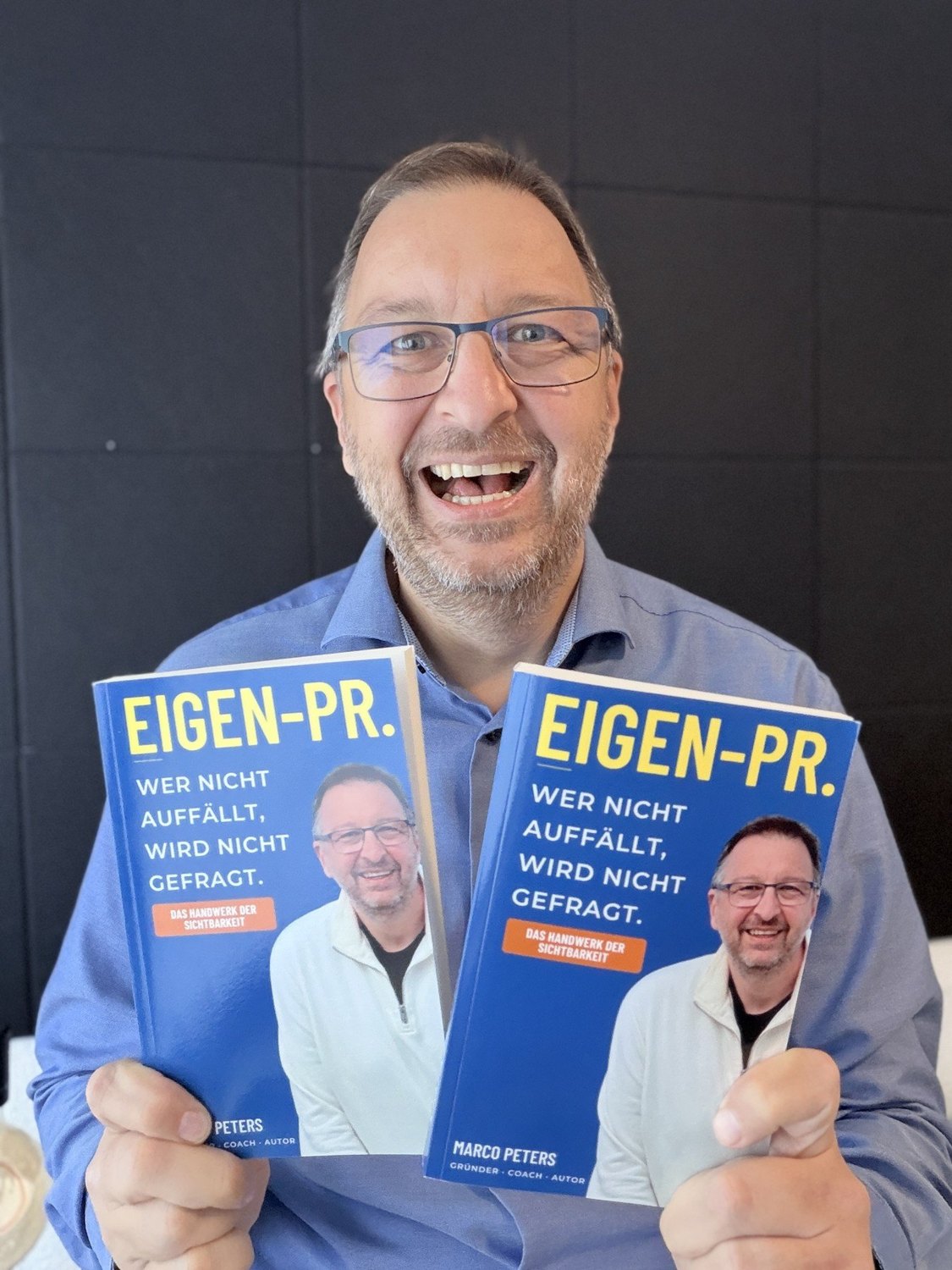 Marco Peters lacht und hält sein Buch EIGEN-PR. in den Händen